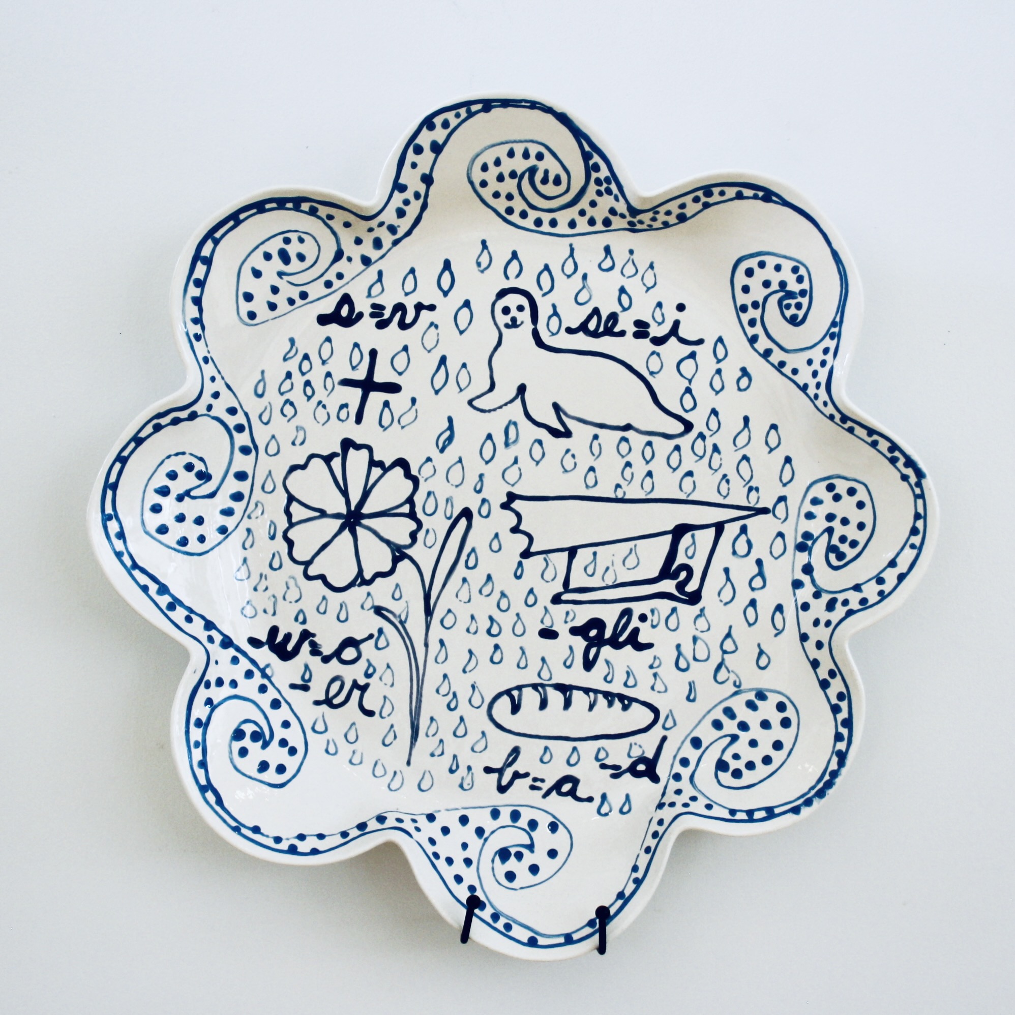 How to Read: Fig 4. Pluvial Flooding, 2024   Fibre clay, low-fired glazes 46 x 46 x 7cm  (18 x 18 x 2.7 inches)  Reading: What we see: a back ground of rain drops framed by undulating waves. What we are being asked to decipher – line drawings and letters. Reading left to right, top to bottom. A plus sign, but we are also told (s=v) thus plus becomes pluv; next an image of seal, here (se=i), thus ial.  Our first word? PLUVIAL; next line, a flower, but, w=o and remove - er (floo) then a silhouette of gliding (–gli) thus ding, FLOODING; Finally, a loaf of bread, however, if  b=a and we are removing -d we have AREA. We are being warned that this is a Pluvial Flooding Area!
